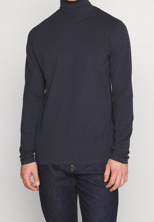 Man wearing a fitted plain black turtleneck with long sleeves and dark blue jeans, standing against a light background.