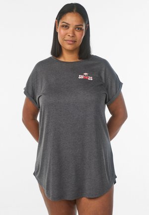 Kvinde med mellemtonet hud og lige sort hår iført en mørkegrå T-shirt med opkørte ærmer og teksten "SNOOZE" samt et kirsebærlogo.