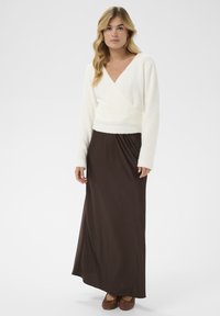 Weißer, flauschiger Wickelpullover mit langen Ärmeln, kombiniert mit einem fließenden braunen Maxirock. Braune geflochtene Schuhe runden das Outfit ab.