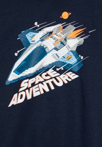Maglietta con una grafica di un'astronave bianca e blu con dettagli arancioni e fiamme, su uno sfondo blu navy. Il testo recita "AVVENTURA NELLO SPAZIO."