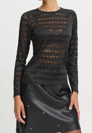 Mujer que lleva una blusa de encaje negra de manga larga y una falda de cuero negra con pequeñas tachuelas plateadas, con las manos descansando cerca de los bordes de la falda.