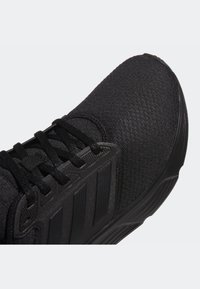 Zapatilla deportiva negra con un upper texturizado, cordones planos y tres rayas laterales distintivas. La suela es curva y tiene un dibujo prominente.