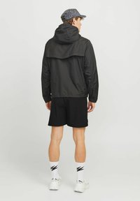 Junger Mann in einer schwarzen Kapuzenjacke, schwarzen Shorts, weißen Turnschuhen, weißen Socken mit schwarzen Zickzackmustern und einer grauen gemusterten Mütze, der steht.