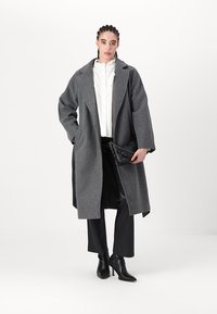 Langer grauer Wollmantel mit übergroßer Passform, mit Reverskragen und seitlichen Taschen, kombiniert mit schwarzen Hosen und schwarzen, hochhackigen Stiefeletten.
