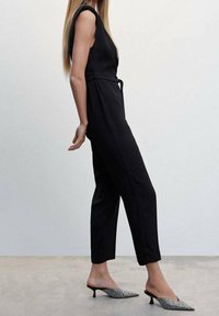 Černý overal s přiléhavým korzetem, bezrukávovým designem a páskem v oblasti pasu; spárovaný s muly s úzkou špičkou v černo-bílé vzorované kombinaci.