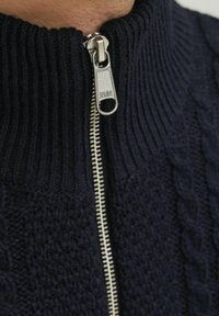 Marineblauer Strickpullover mit hohem Rippkragen und silbernem Reißverschluss. Verfügt über ein strukturiertes Kabelstrickmuster und glattes Innenfutter.