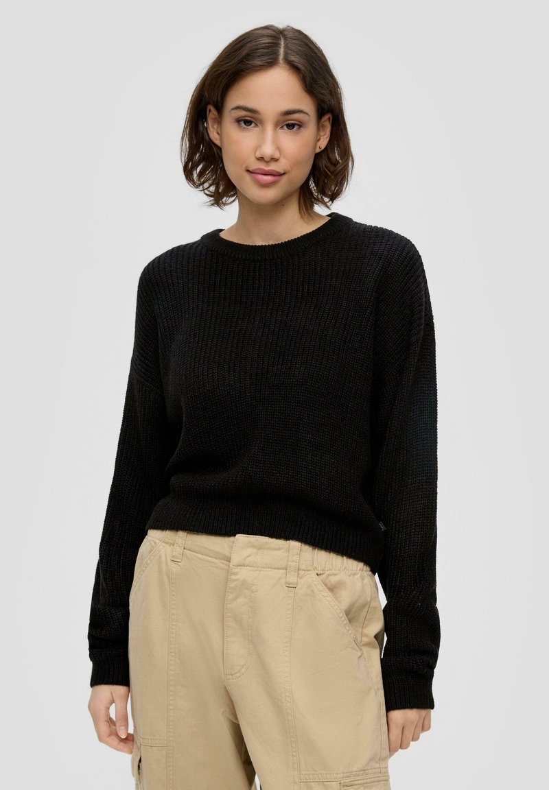Schwarzer Strickpullover mit Rundhalsausschnitt und langen Ärmeln, kombiniert mit beigen Cargo-Hosen mit elastischem Bund und seitlichen Taschen.