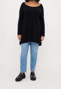 Persoon in een zwarte langwerpige tuniek, lichtblauwe jeans, zwarte platvorm loafers met studs, en een parelketting, staand tegen een effen achtergrond.