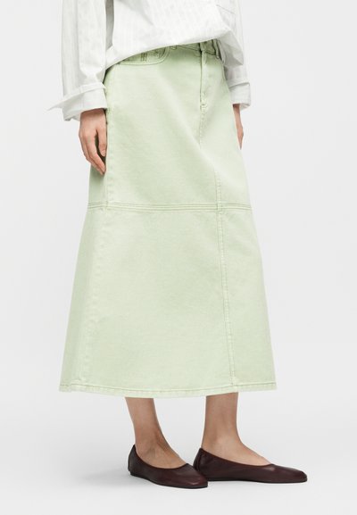 Femme portant une jupe midi vert pâle, un chemisier blanc à manches longues et des chaussures plates marron foncé, debout devant un fond blanc uni.