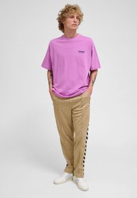 Lyse lilla T-shirt med korte ærmer og et logo, parret med beige bukser med et subtilt mønster og sorte striber. Hvide sneakers.