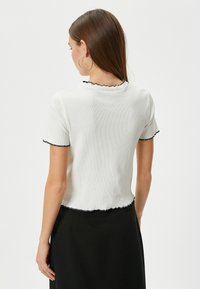 Hvid ribbet kortærmet top med sorte flæser langs udskæringen og ærmerne, der har et cropped design og tekstureret stof.