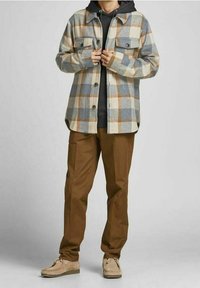 Camicia di flanella a quadri nei toni beige, blu e arancione, con un sottogiacca nera con cappuccio sotto. I pantaloni marroni e le scarpe beige chiare completano l'outfit.