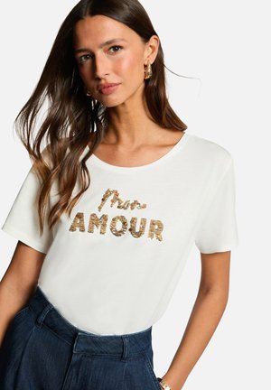 T-shirt blanc à manches courtes avec le texte en sequins dorés "Moi, Amour" sur le devant. Le tissu semble lisse et léger.