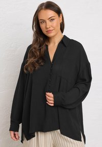 Camicia oversize nera con colletto, taschino frontale e maniche lunghe, realizzata in un tessuto morbido e leggero.
