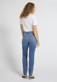 Käharate juustega naine, kes kannab valget lühikeste varrukatega särki, siniseid kitsaid teksapükse ja pruune kontsakingi, seistes seljaga vaataja poole.