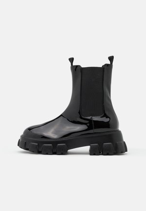 Bottine en cuir noir brillant avec semelle épaisse à crampons et panneaux latéraux élastiques sur un fond clair uni.