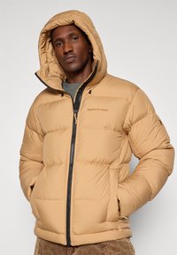 Bež puffer jakna sa zip zatvaranjem, kapuljačom i elastičnim manžetama. Ima kutijasti kviltani dizajn i Peak Performance logotip na grudima.