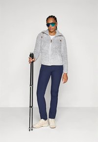 Pilkas flisinis džemperis su užtrauktuku ir gaubtu, tamsiai mėlynos kelnės ir balti sportbačiai; asmuo laiko juodą žygio lazdą ir dėvi mėlynas saulės akinius.