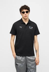 Νεαρός άνδρας με σγουρά μαλλιά, φοράει μαύρο πόλο Puma BMW Motorsport και γκρι σορτς, στέκεται με τα χέρια στις τσέπες μπροστά σε απλό φόντο.