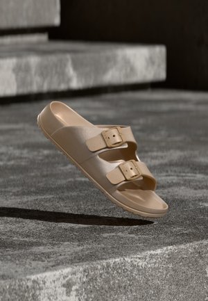 Yksi beige liukusandaali, jossa on kaksi säädettävää solkiremmiä, lepää kivisten askelmien päällä pehmeässä valaistuksessa.