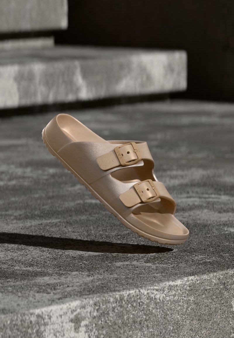 Μονό μπεζ σανδάλι slide με δύο ρυθμιζόμενα λουριά με αγκράφες που ακουμπούν σε πέτρινα σκαλοπάτια με απαλό φωτισμό.
