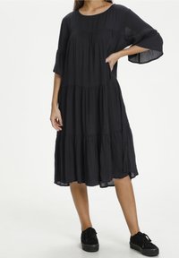Mujer con un vestido midi negro suelto y con capas, de mangas hasta el codo, y zapatillas de plataforma negras, de pie con una mano en la cadera.