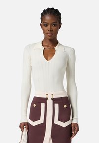 Top bianco a maniche lunghe a coste con scollo a V e dettaglio in catena metallica sul colletto; abbinato a una gonna marrone con dettagli bianchi sulle tasche.