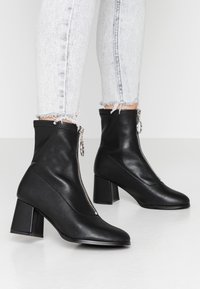 Bottines en cuir vegan noir avec fermeture éclair, talons carrés et texture lisse. Présente un design épuré avec des accents minimalistes.