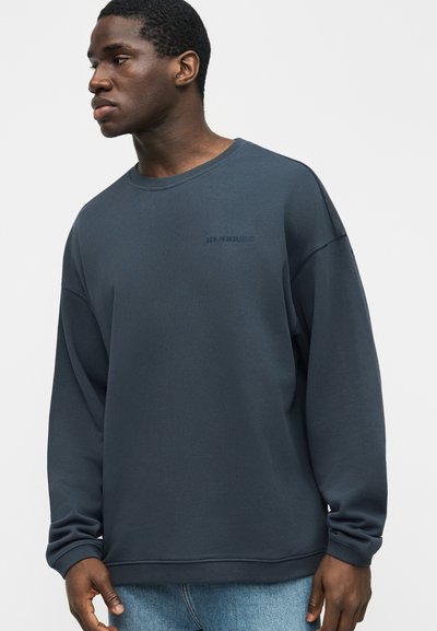 Νέος άνδρας με σκουρό μπλε φαρδιά φούτερ και ανοιχτό μπλε τζιν, κοιτώντας προς τα αριστερά μπροστά σε μονόχρωμο ανοιχτό φόντο.