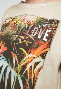 Jasno beżowa bawełniana koszulka z kolorową grafiką roślinną i napisem "SUNSET LOVE" w srebrnych literach oprawionym w ciemną ramkę.