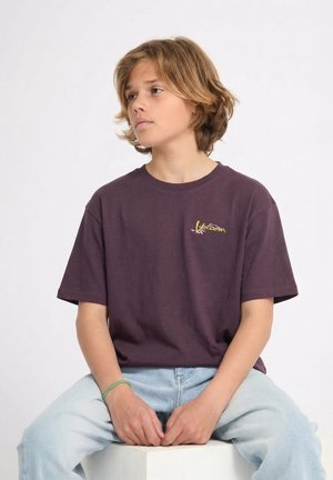 Adolescent aux cheveux blonds mi-longs portant un T-shirt Volcom violet foncé et un jean bleu clair, assis les mains jointes.