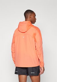 Veste légère à capuche de couleur orange vif, avec une texture lisse et un accent réfléchissant à l'arrière. Conçue pour les activités sportives.