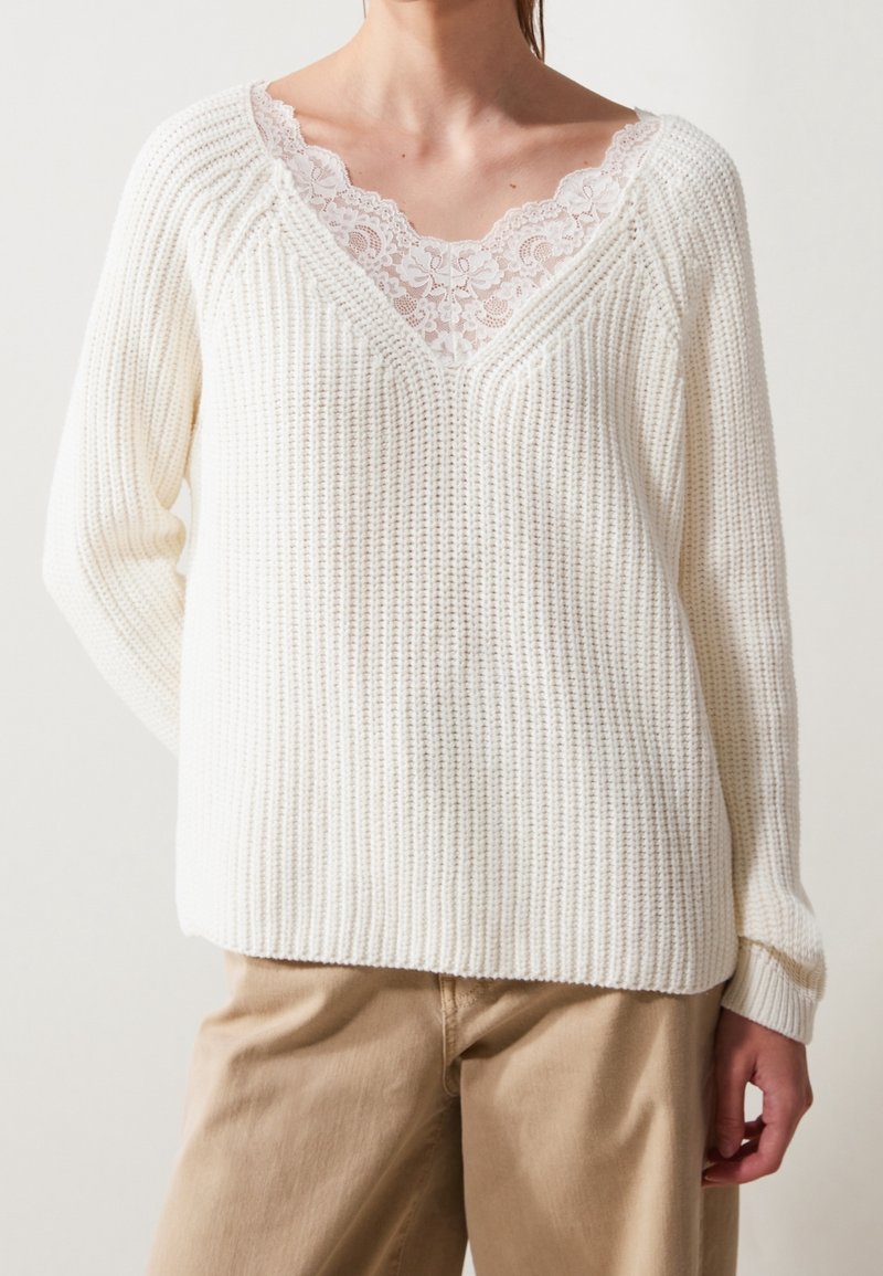 Lehký bílý svetr s texturou, výstřihem do V a krajkovou lemovkou. Má žebrovaný úplet a volnější střih, s dlouhými rukávy a spadlými rameny.