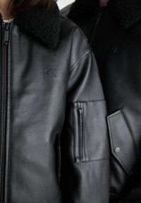 Μαύρο δερμάτινο μπουφάν με φουσκωτό μαύρο γιακά, κλείσιμο με φερμουάρ και πολλαπλές τσέπες με φερμουάρ. Διακριτική λεπτομέρεια λογότυπου στο στήθος.