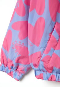 Veste colorée avec un motif de cœurs rose et bleu, fabriquée dans un matériau lisse et léger, dotée de poignets élastiques et d'une poche latérale.
