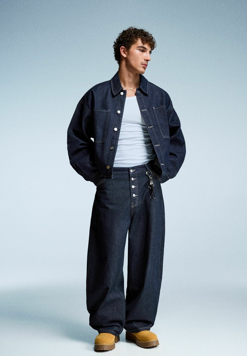 Giacca di jeans blu scuro e pantaloni a gamba larga con chiusura a bottoni e cuciture visibili. Il modello indossa una canotta bianca e calzature color sabbia.