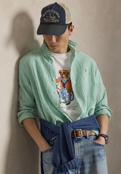 Ung mann som har på seg en grønn stripete skjorte over en t-skjorte med bamsegrafikk, blå strikkegenser knytt rundt livet, jeans og en baseballcaps.