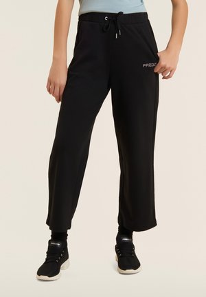 Henkilö, jolla mustat nyörityshousut ja mustat valkopohjaiset tennarit vaalealla taustalla, seisoo käsi taskussa.