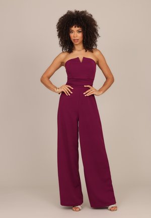 Frau mit lockigem Haar, die einen trägerlosen, pflaumenfarbenen Jumpsuit und silberne High Heels trägt, steht mit den Händen in der Hüfte vor einem schlichten Hintergrund.