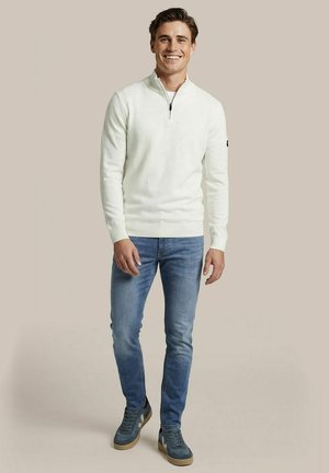 Man die staat en lacht, gekleed in een witte trui met een kwart ritssluiting, blauwe spijkerbroek en blauwe sneakers tegen een effen beige achtergrond.