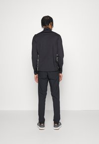Zwarte sportieve zip-up jacket met hoge kraag, gecombineerd met bijpassende zwarte broek en grijze sneakers, getoond vanuit de achterkant.