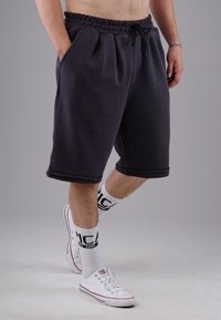 Dunkelgraue Baumwollshorts mit elastischem Bund, Kordelzug, Seitentaschen und umgeschlagenem Saum, kombiniert mit weißen Sneakers und Socken.