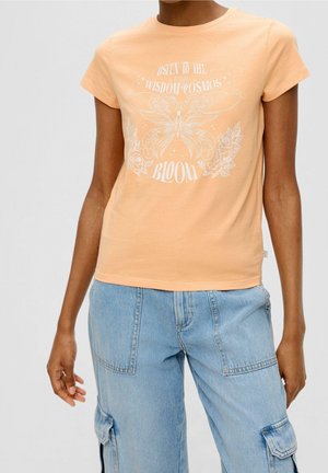 Osoba nosząca brzoskwiniowy t-shirt z motywem motyla i kwiatów oraz tekstem "Listen to the wisdom cosmos bloom", połączony z jasnoniebieskimi spodniami typu cargo.