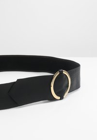 Svart lærbelte med en sirkulær spenne i gulltone. Designet inkluderer en flat stropp og en slank finish.