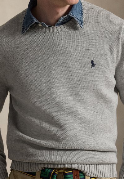Man draagt een grijze gebreide trui over een blauw denimshirt met een gestreepte riem en kaki broek, bij de nek en taille bijgesneden.