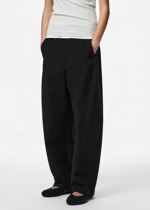 Personne portant un pantalon noir ample, des chaussures noires sans lacets et un haut blanc sans manches, debout les mains dans les poches sur fond blanc.