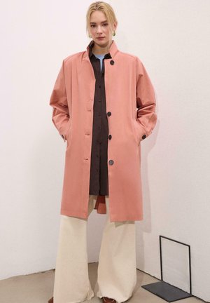 Plavuša stoji naslonjena na bijeli zid noseći labavi rozi kaput preko tamne majice sa širokim kremasto obojenim hlačama i smeđim cipelama.