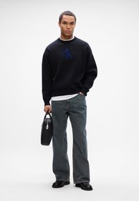 Čierna mikina s modrým logom "CK", kombinovaná s voľnými tmavými džínsami a čiernymi topánkami. Drží čiernu tašku s rúčkou.