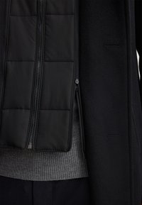 Cappotto nero con un rivestimento interno trapuntato, dotato di cerniere su entrambi gli strati, un maglione a maglia grigia testurizzata sotto e un tessuto esterno liscio.