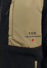 Giacca in pile con una sezione inferiore nera e testurizzata e una parte superiore liscia color tan, caratterizzata dalla scritta "R.D.D. ROYAL DENIM DIVISION" stampata in nero e un punto rosso.